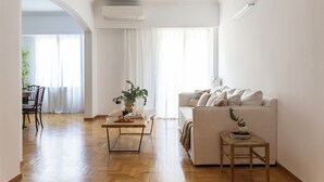 Uma sala de estar moderna com piso de madeira, um sofá branco, uma mesinha com uma planta e um espaço de jantar com uma cadeira e mesa.