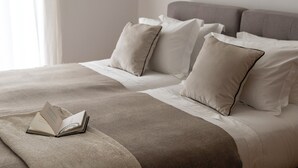 Cama arrumada com um cobertor bege e almofadas, um livro sobre a cama e uma janela com cortinas.