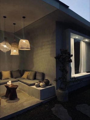 Un área de estar moderna al aire libre con sofá, cojines y una mesa, iluminada con lámparas colgantes.