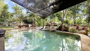 Ein klares Schwimmbecken mit steiniger Kante, umgeben von Bäumen und einem Metallzaun.