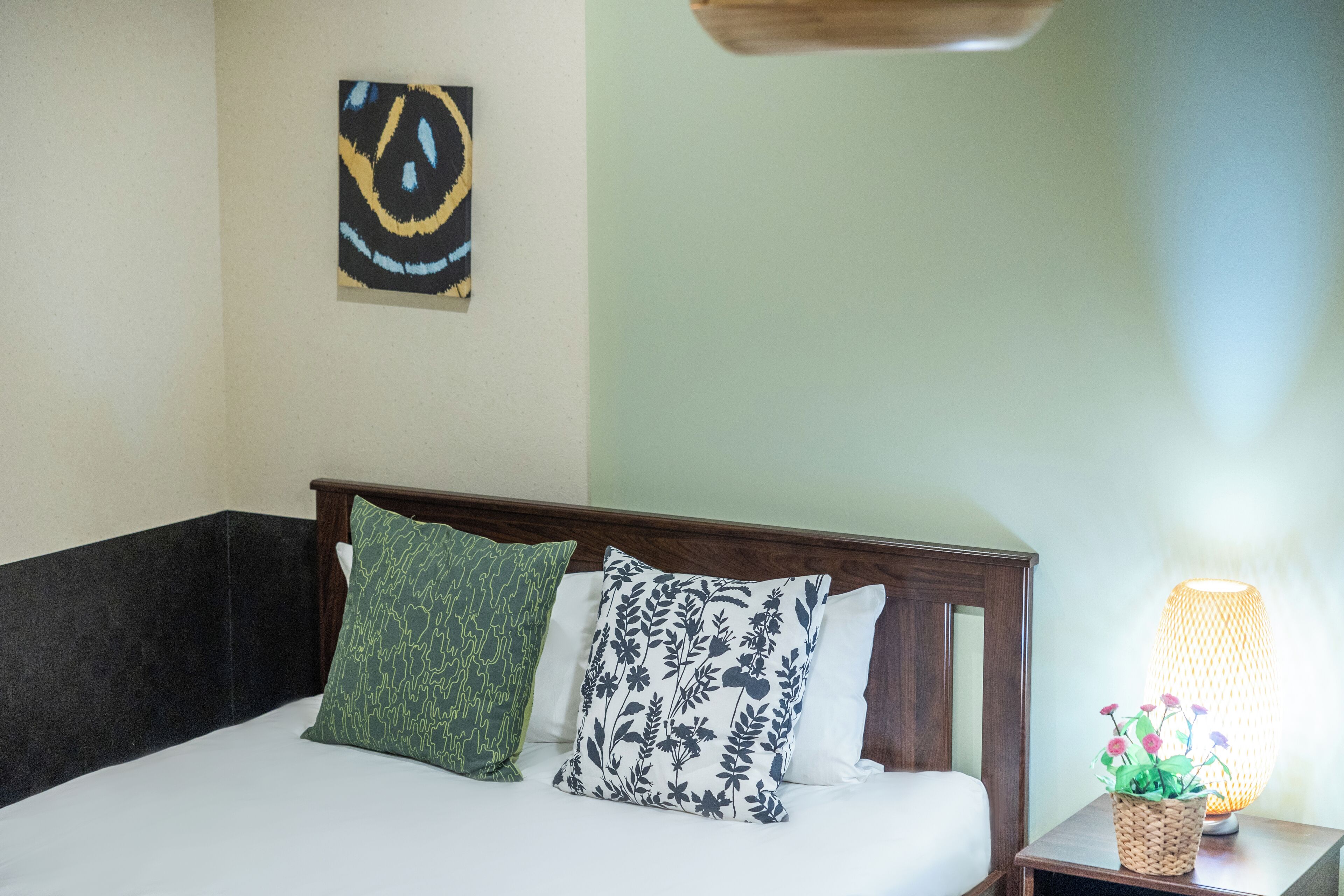 A neatly made bed with two pillows, a small bedside table with a lamp and a basket, and a wall art piece above the bed.