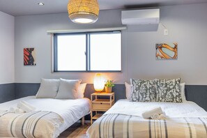 A hotel room with two beds, a window, a wall-mounted air conditioner, and a small table with a lamp and a plant.
