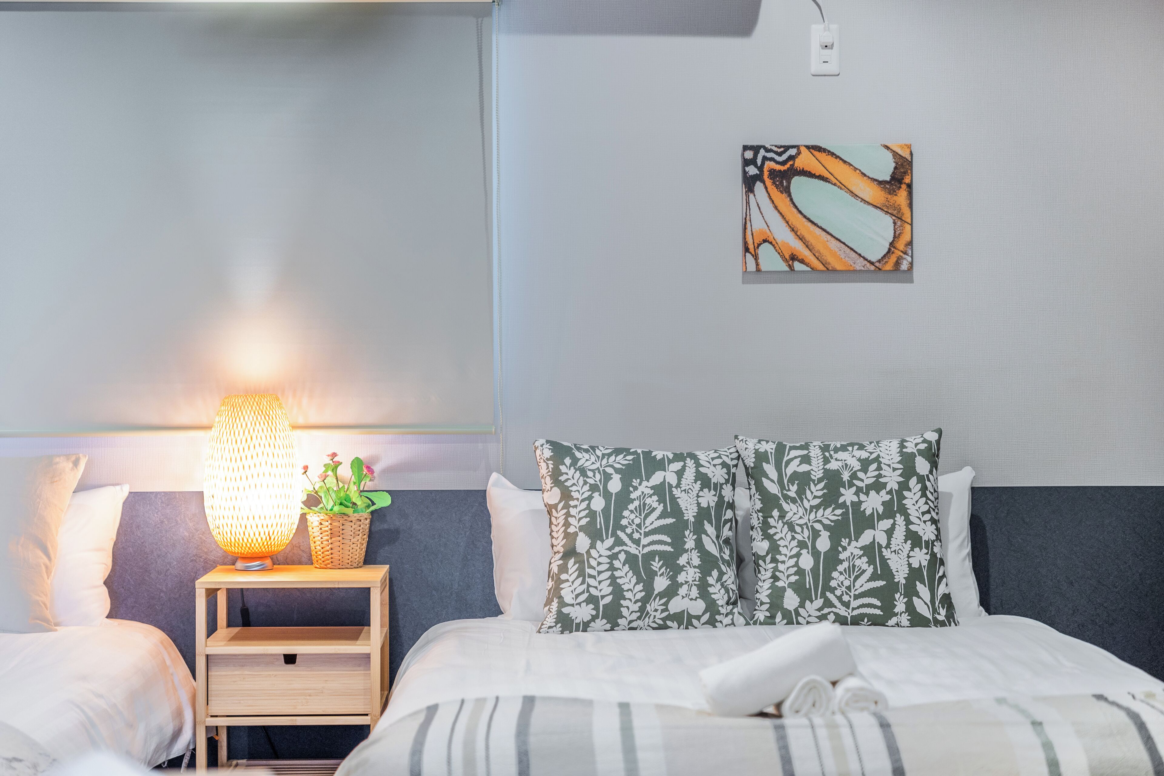 Un lit bien fait, recouvert d’une couvre-lit à rayures, une lampe de chevet, une petite table de nuit en bois ornée d’une plante, et un tableau abstrait accroché au mur.