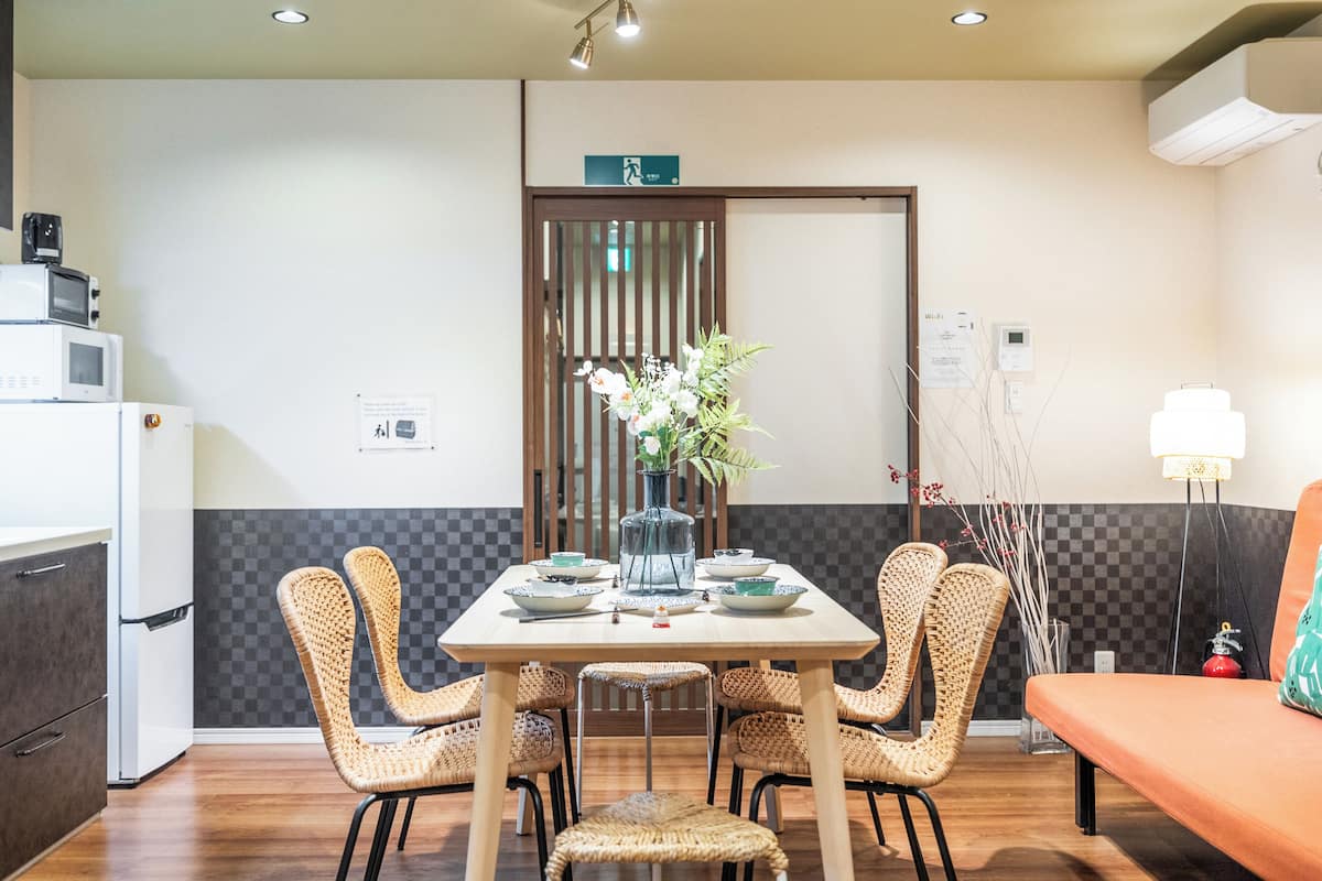 A dining area with a table set for four, surrounded by wicker chairs, a vase with flowers, and a wooden door in the background.