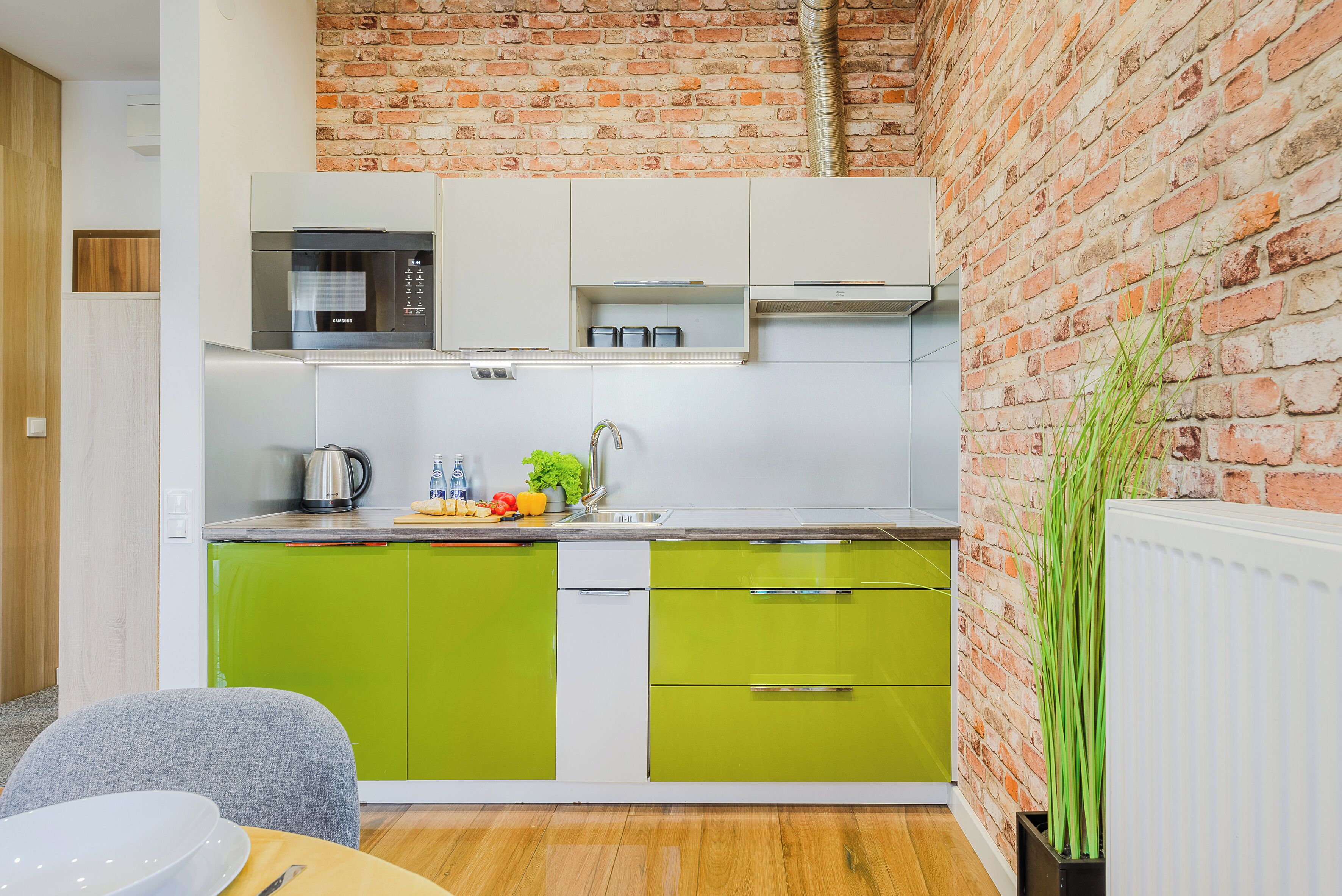A modern kitchen with green and white cabinets, a stainless steel microwave, and a brick wall.