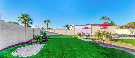 A well-maintained artificial green lawn with a white wall, palm trees, and red umbrellas.