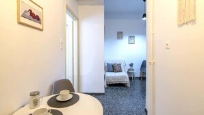 A small, cozy room with a dining table set for two, a bed with a white cover, and a small side table with a lamp. - Your perfect choice for stay in Athens (Athina)