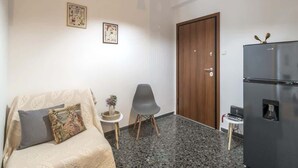 A small, well-lit room with a sofa, a chair, a small table with a vase, a refrigerator, and a wooden door. - Your perfect choice for stay in Athens (Athina)