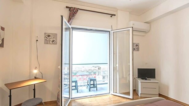 A bedroom with a bed, a desk with a chair, a TV, and a sliding glass door leading to a balcony with outdoor furniture. - Your perfect choice for stay in Athens (Athina)