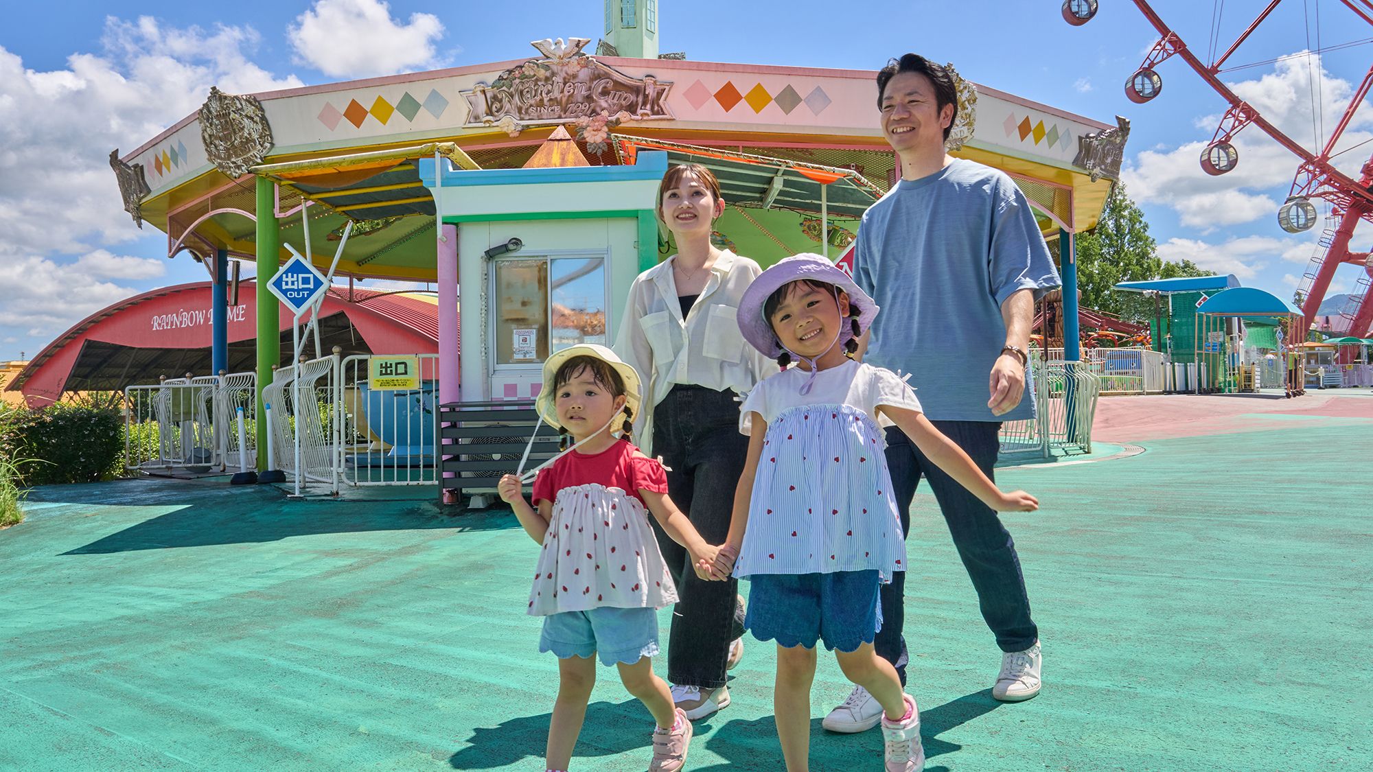 Eine Familie steht vor einer bunten Attraktion in einem Freizeitpark, im Hintergrund ist ein Riesenrad zu sehen.