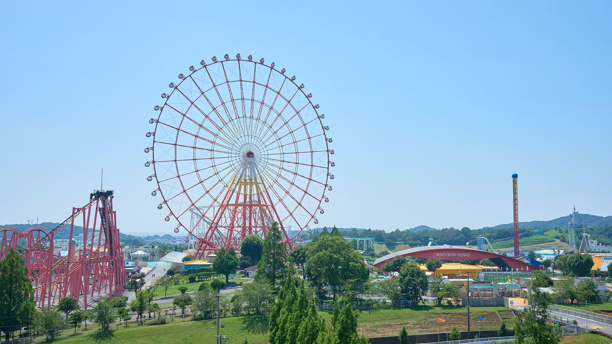Ein großes Riesenrad, eine Achterbahn und ein Gebäude mit der Aufschrift 'Rainbow Dome' in einer Parkanlage.