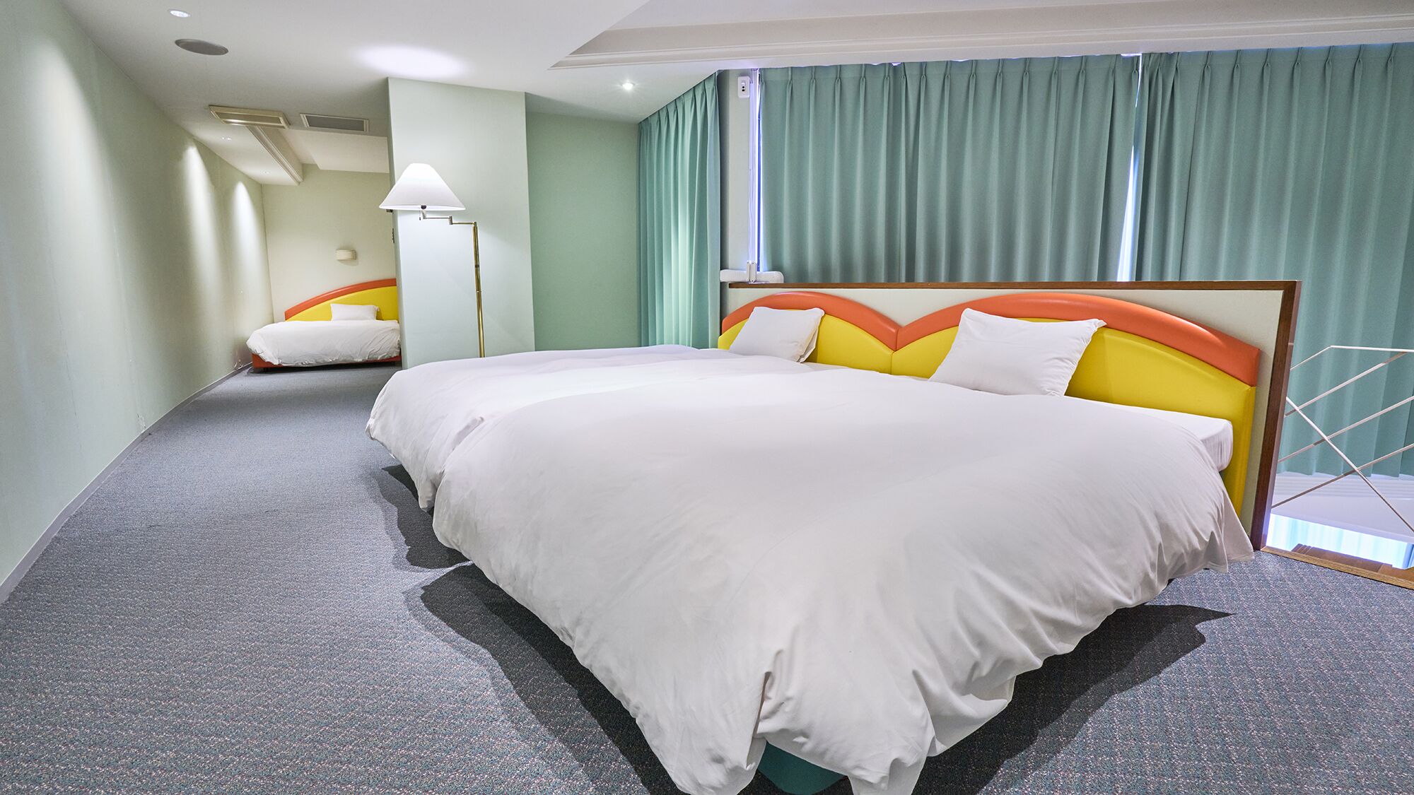 Habitación de hotel con una cama grande, una cama más pequeña, una mesita de noche, una lámpara y vistas al exterior a través de una ventana con cortinas.