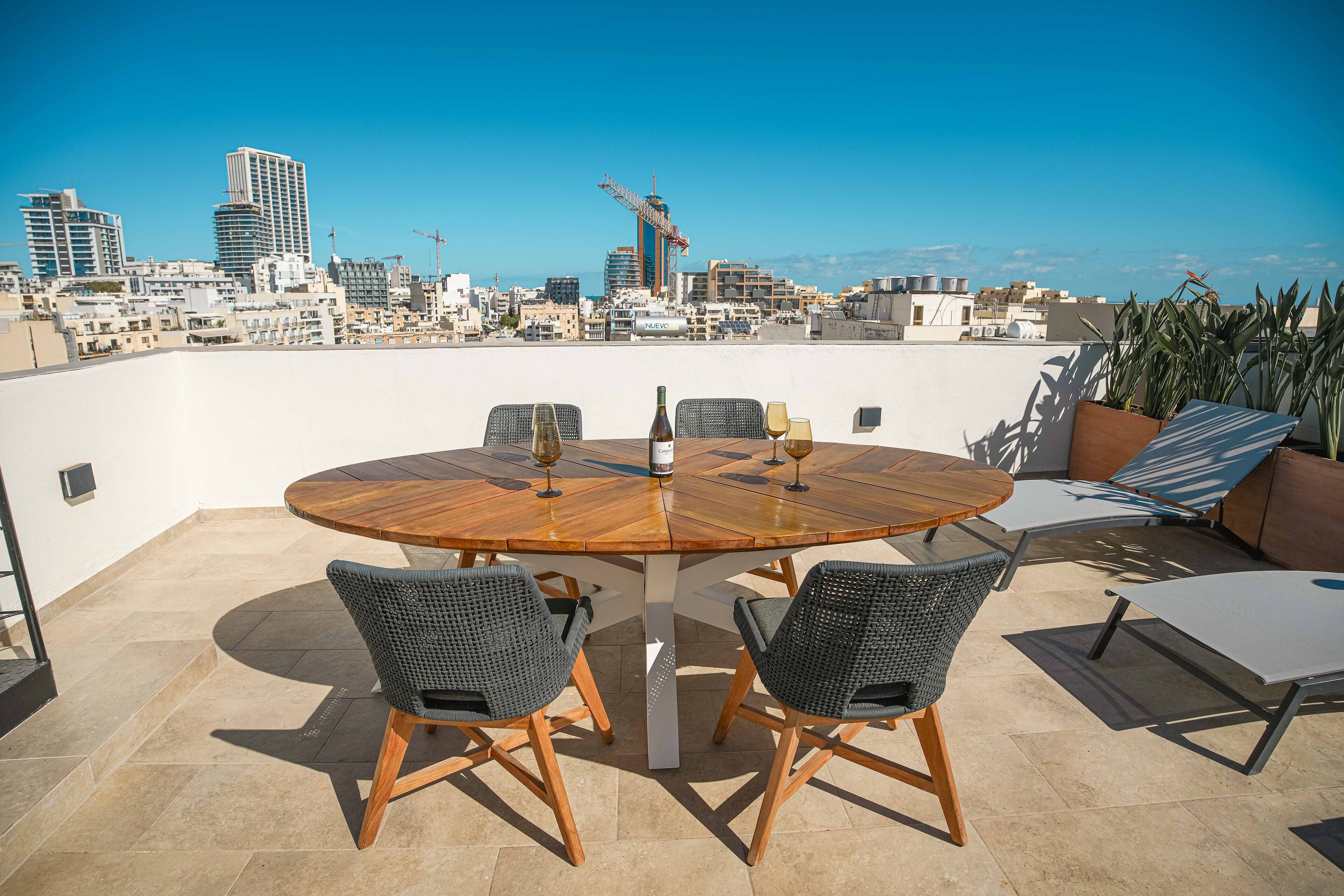 Terraza en la azotea con una mesa de madera redonda y cuatro sillas de mimbre, con vistas a un paisaje urbano con edificios modernos y una grúa de construcción.