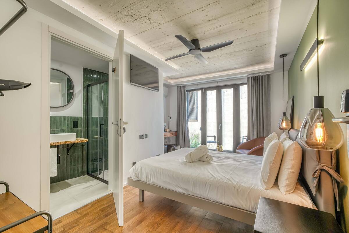 Egy modern hálószoba, amelyben található egy ágy, éjjeli lámpák, egy mennyezeti ventilátor, valamint egy fürdőszoba zuhanyzóval és mosdókagylóval.