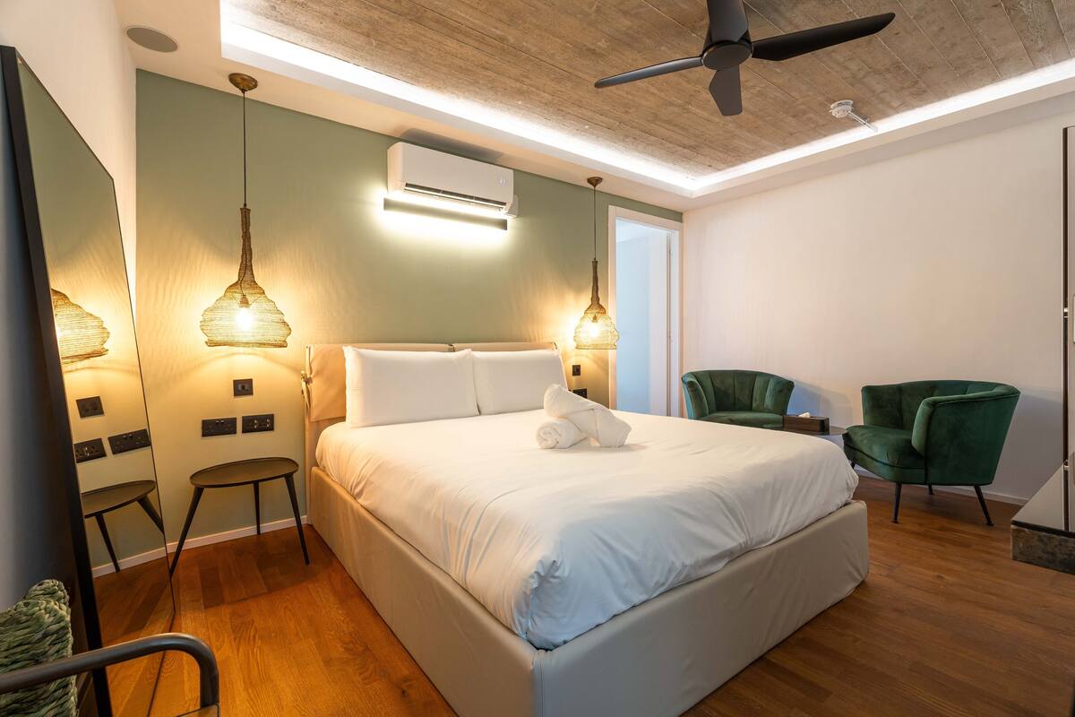 Egy modern szállodai szoba, amelyben egy nagy ágy, két éjjeliszemélyzet, egy zöld karosszék és egy mennyezeti ventilátor található.