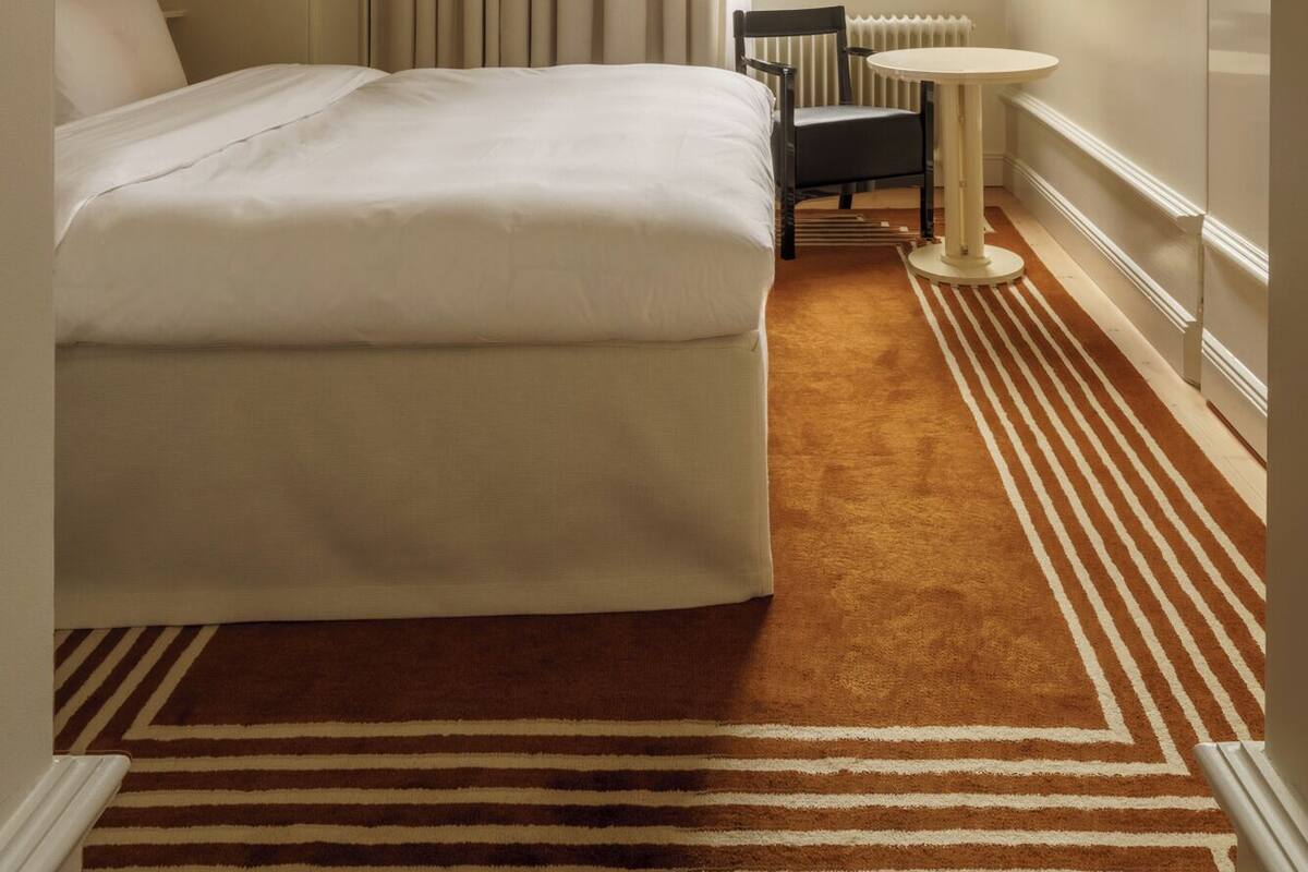 Habitación de hotel con una cama, una silla, una mesita, un radiador y un televisor en la pared.