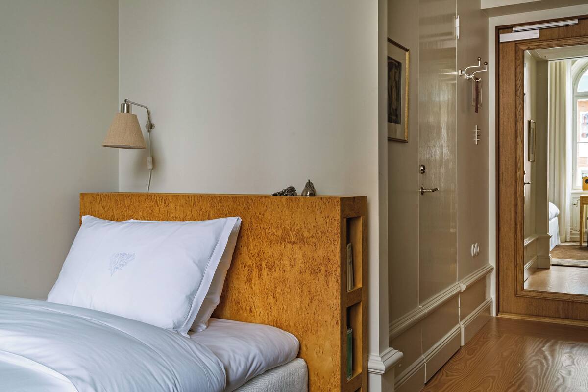 Un dormitorio con cabecera de madera, cama con ropa de cama blanca, lámpara de noche, armario y espejo.
