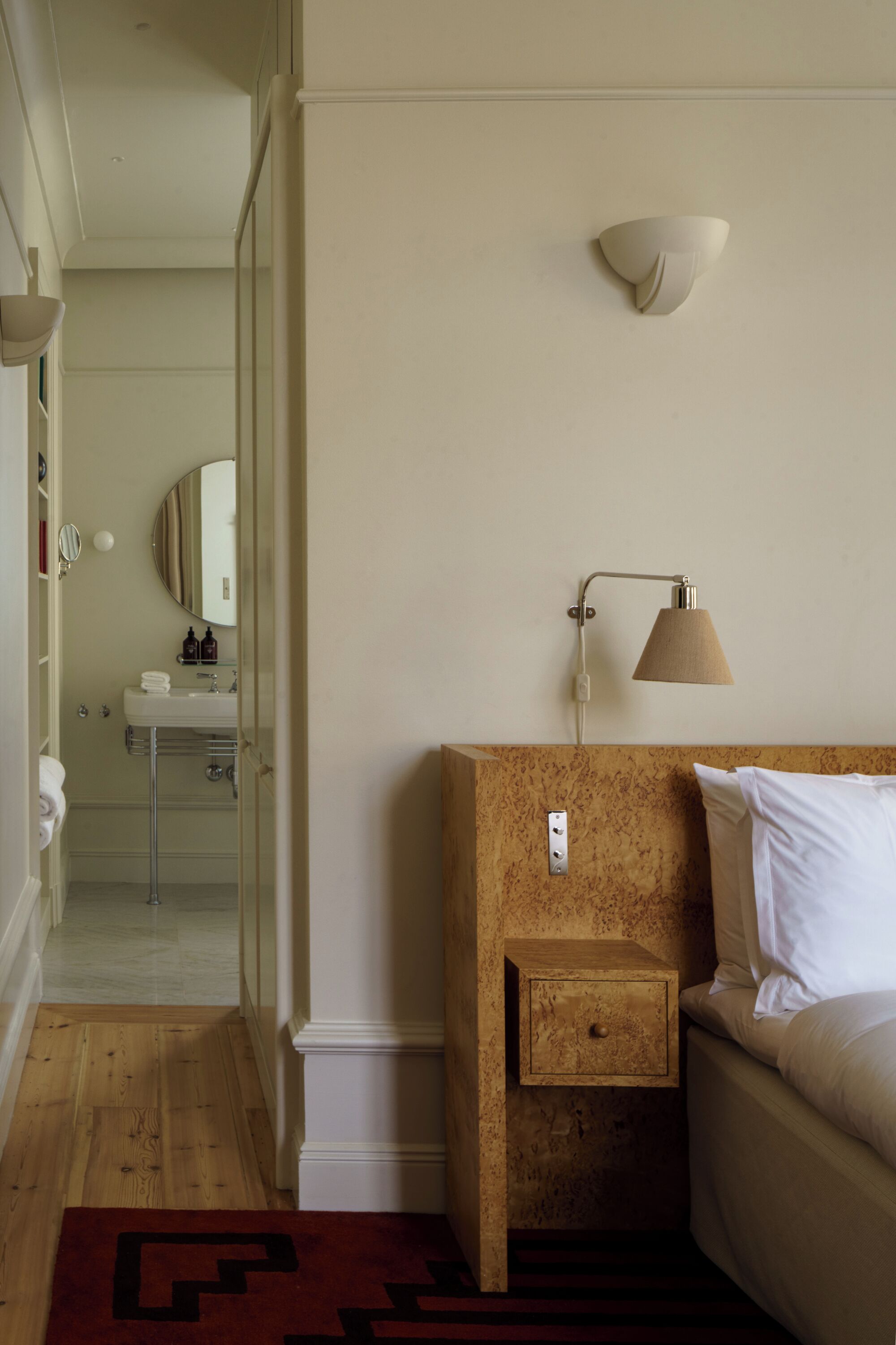 Ett hotellrum med en sänggavel i trä, en sänglampa, ett nattduksbord, en säng, en liten låda, en spegel och ett badrum som syns genom en öppen dörr.