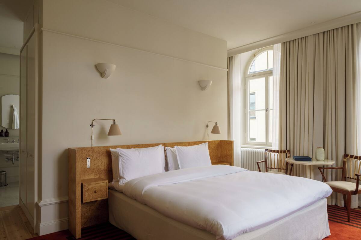 Habitación de hotel con una cama grande, mesitas de noche, una pequeña mesa con sillas y una ventana con cortinas.