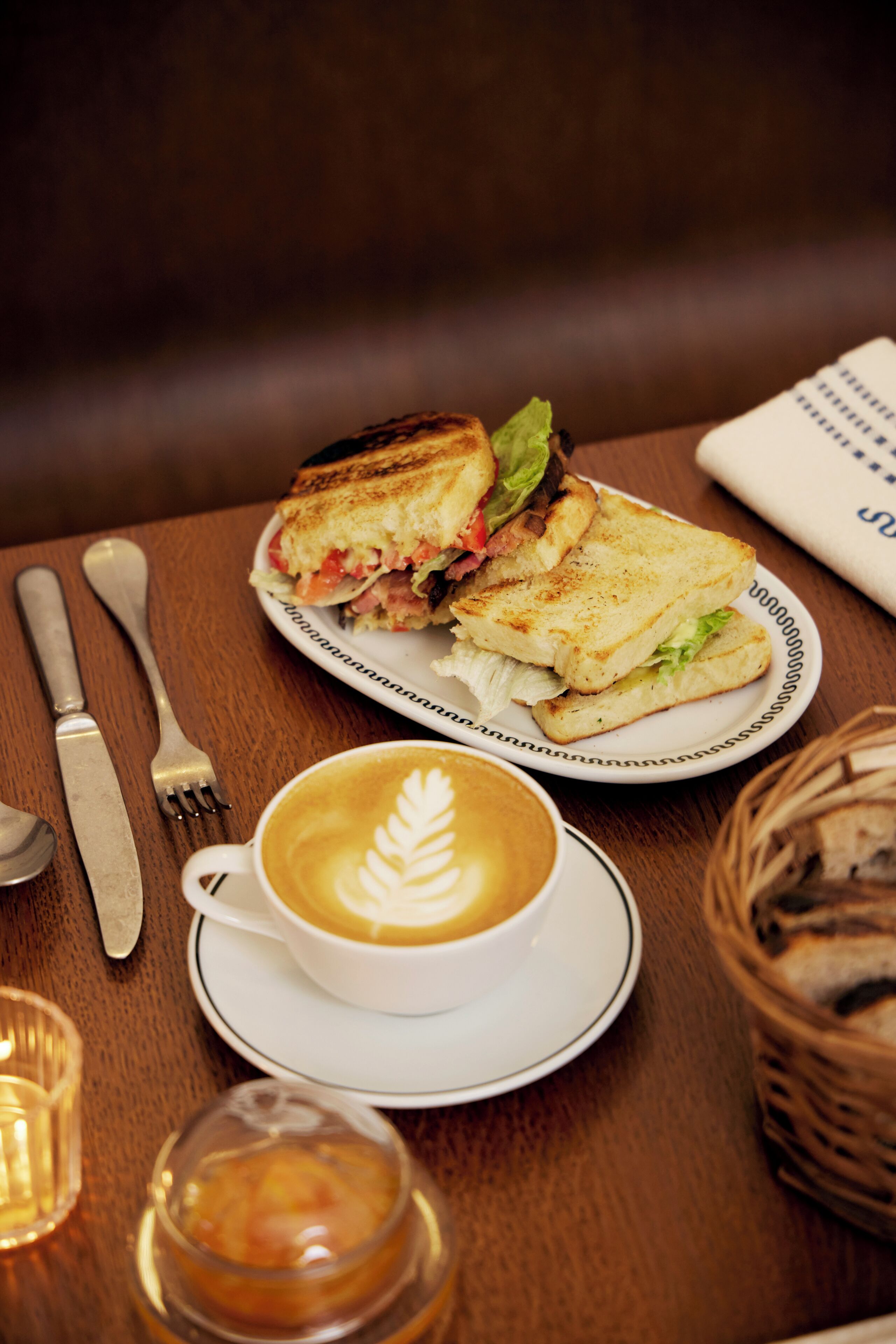 A sandwich on a plate, a cup of coffee with latte art, a fork, a knife, and a candle on a wooden table.