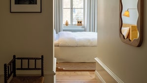 Una habitación de hotel con suelo de madera, cama con ropa de cama blanca, ventana con cortinas, un cuadro enmarcado en la pared y un espejo con lámpara.