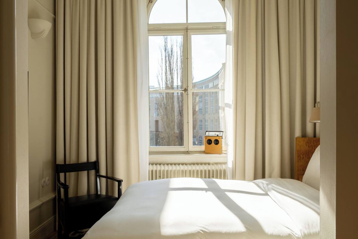 Una habitación de hotel con una cama grande, una ventana con cortinas, una silla y un radiador.