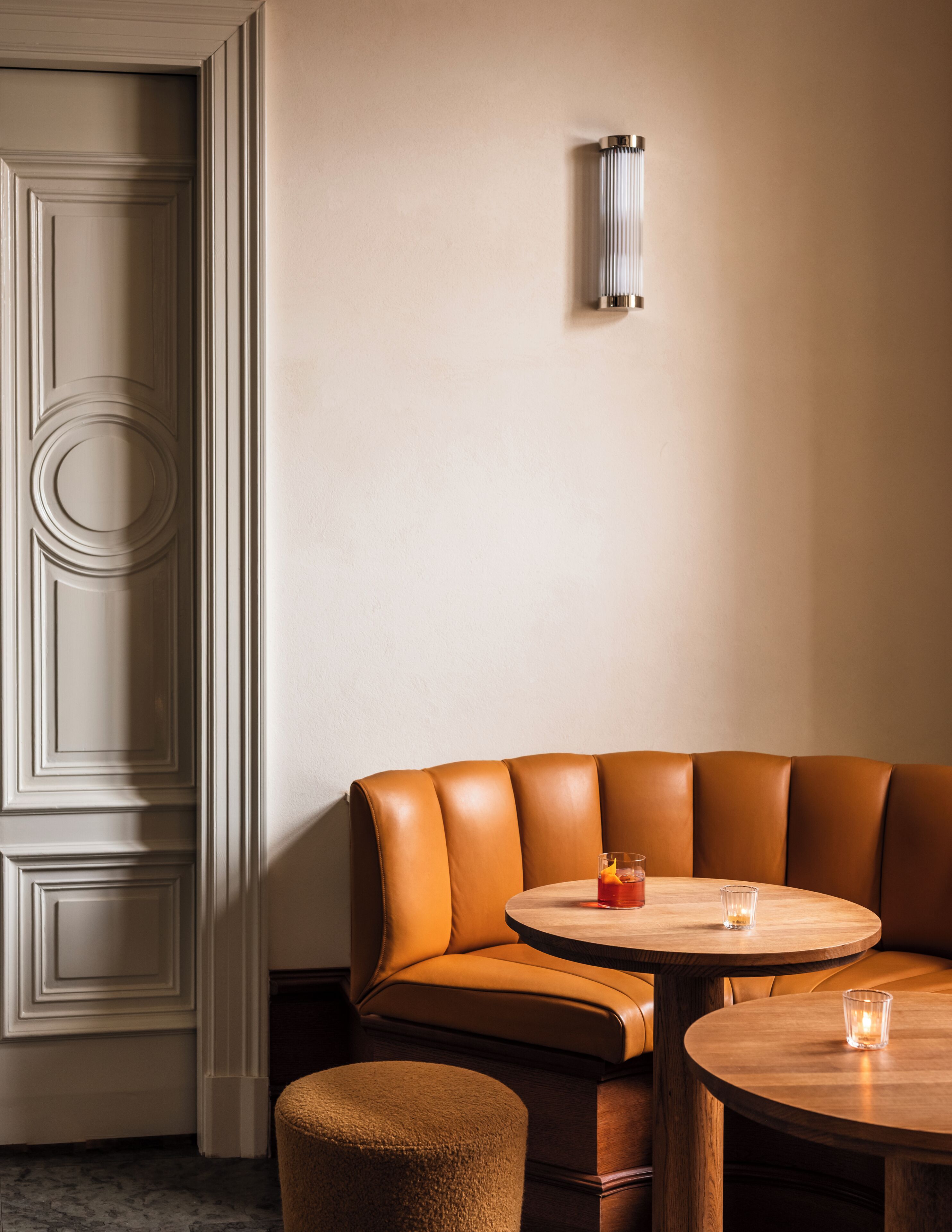 A corner seating area with a cushioned bench and a round wooden table, a wall-mounted light fixture, and a door with decorative paneling.