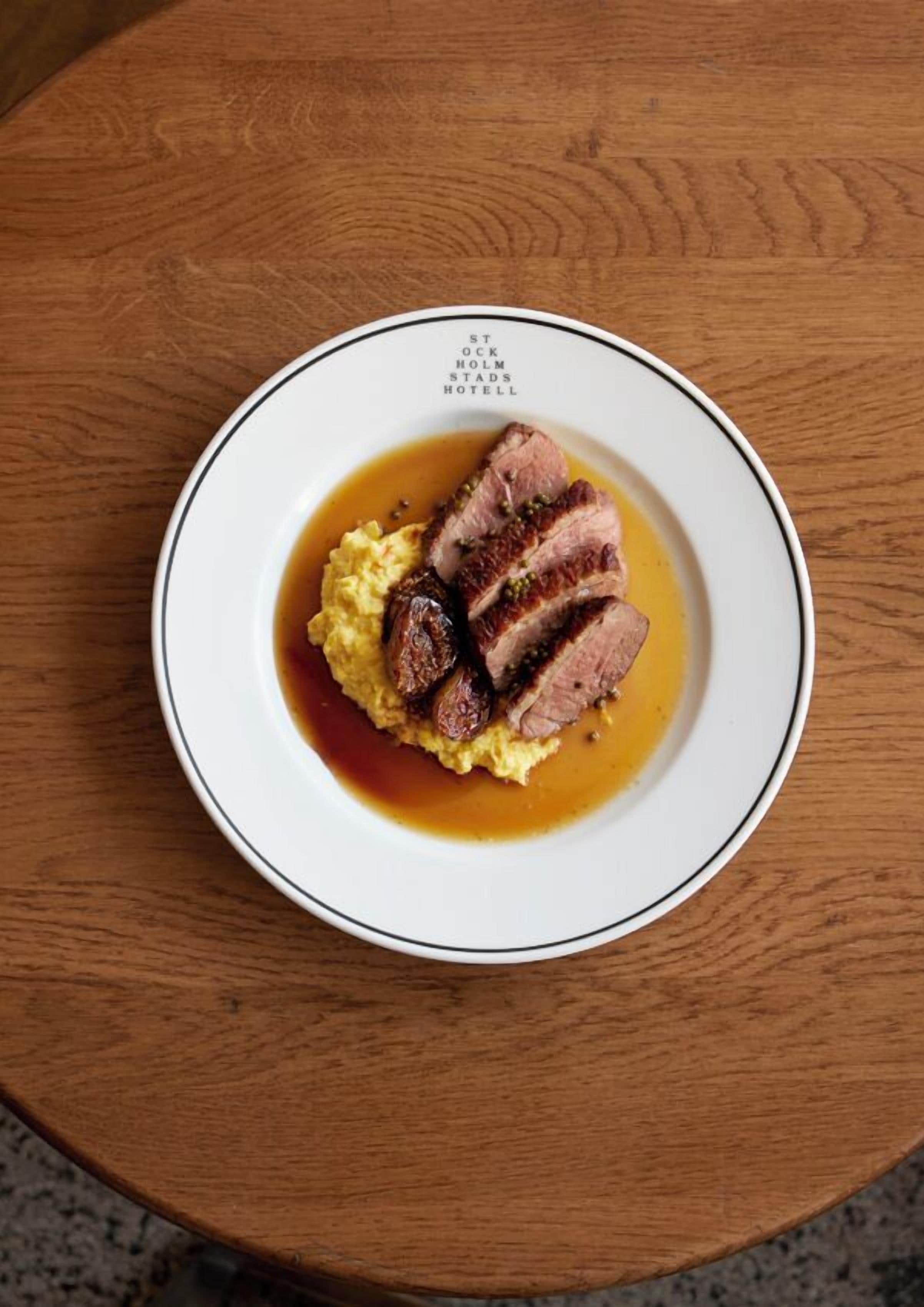 Een bord eten met een witte rand en zwarte rand, met plakjes vlees en aardappelpuree met bruine saus.