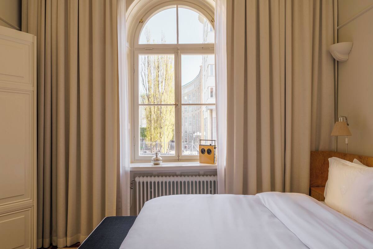 Una habitación de hotel con un ventanal grande, un radiador, una cama con ropa blanca, un cabecero de madera y una lámpara de noche.