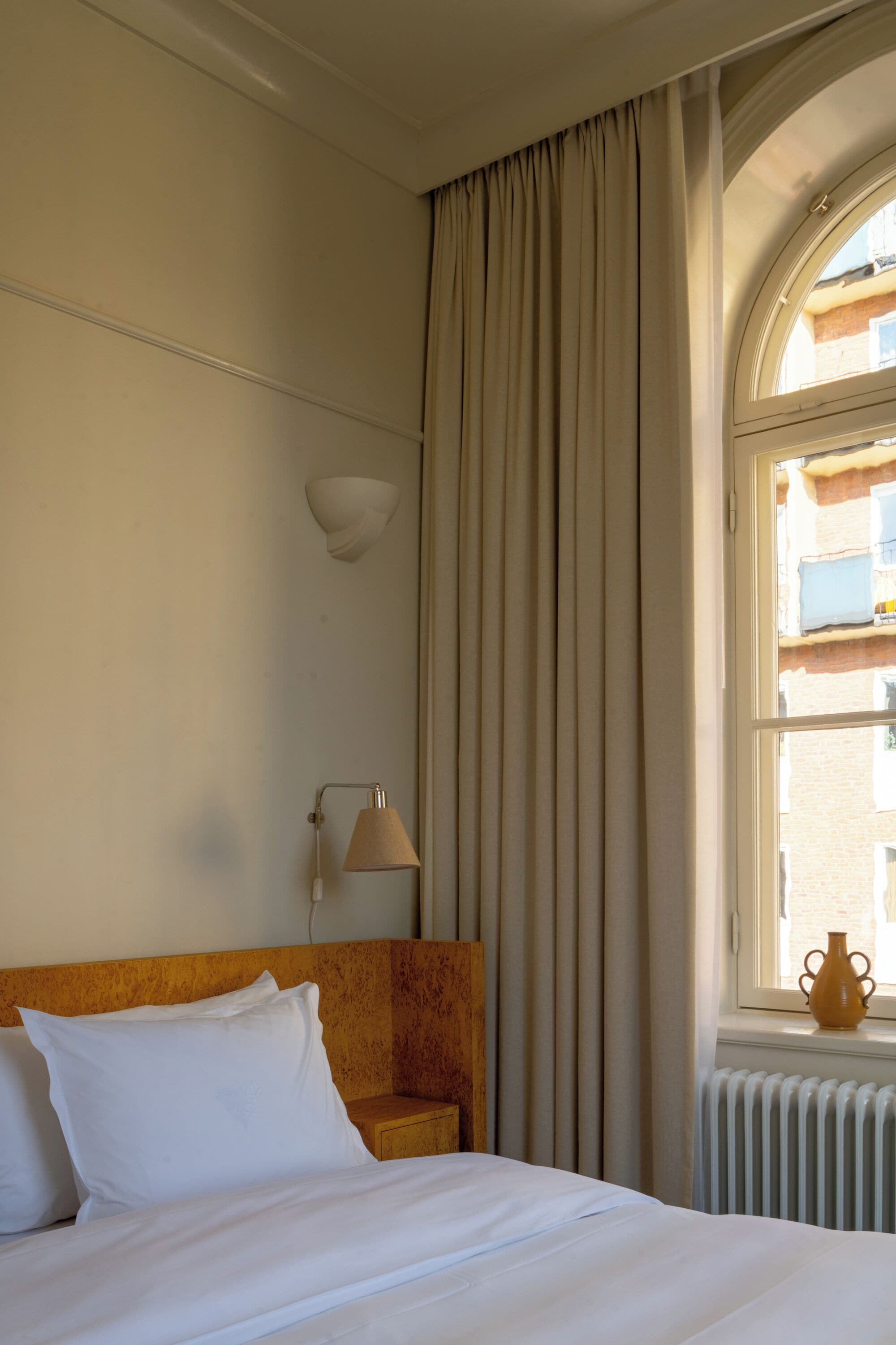 Quarto de hotel com cama, mesa de cabeceira, luminária, janela com cortinas e radiador.