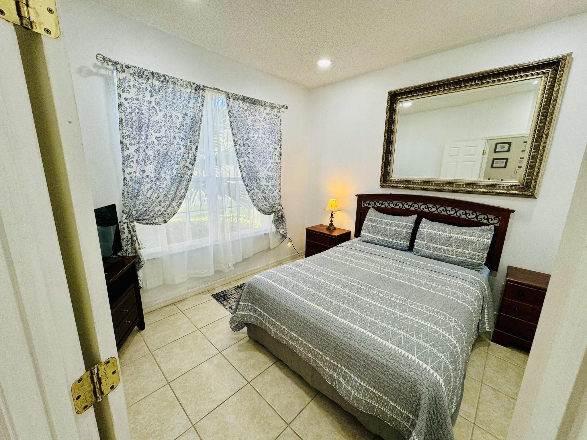 Een slaapkamer met een bed, gordijnen, een nachtkastje, een lamp, een spiegel en een deur.