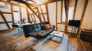 Een loft-achtige kamer met zichtbare houten balken, een zwarte leren bank, een salontafel en een televisie.
