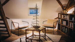 Een gezellig loft slaapkamer met een schuine plafond, een klein raam, een decoratieve boom, twee stoelen, een boekenkast en een houten vloer.