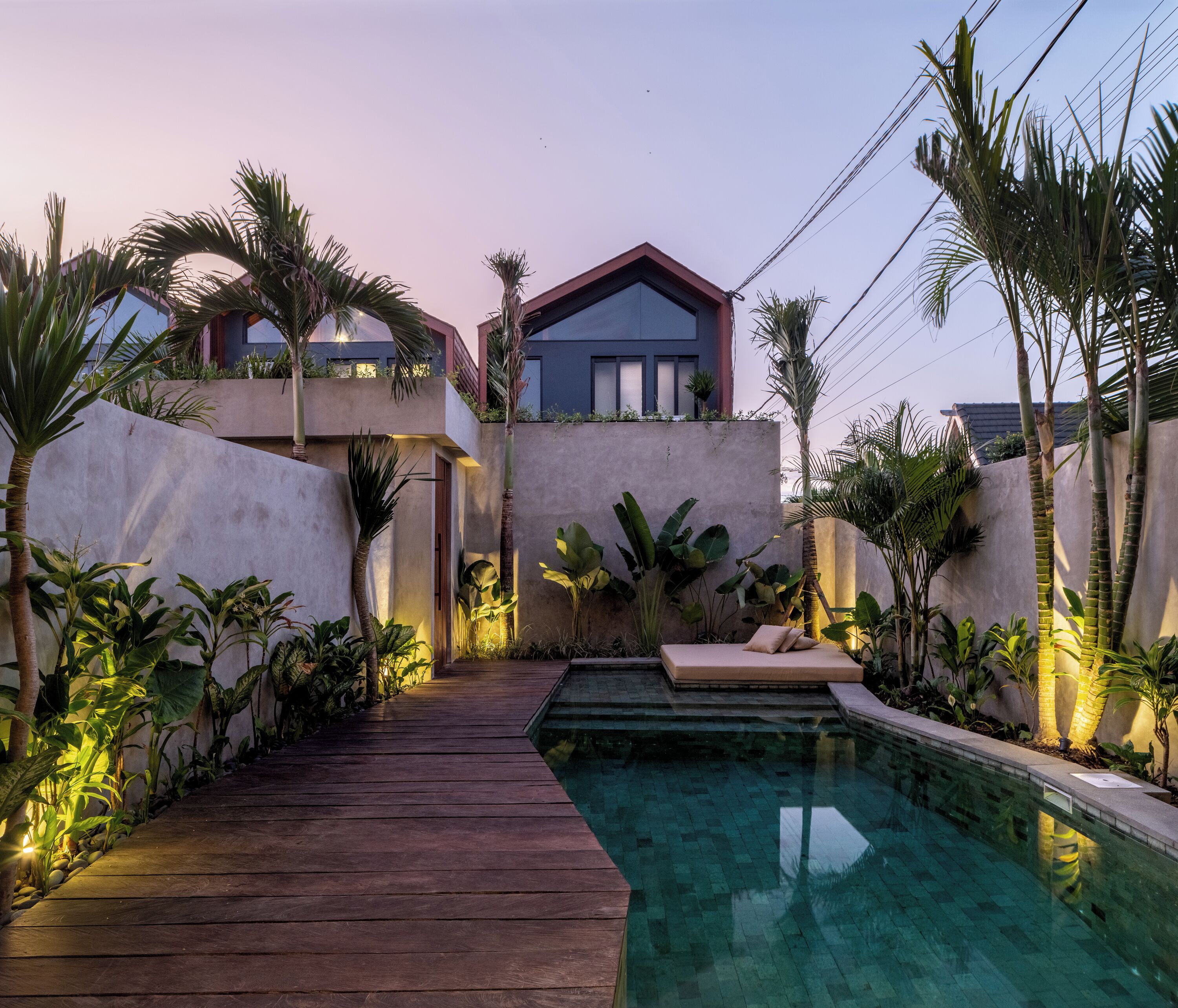 A modern outdoor pool area with a wooden deck, surrounded by lush tropical plants and a two-story building with large windows.