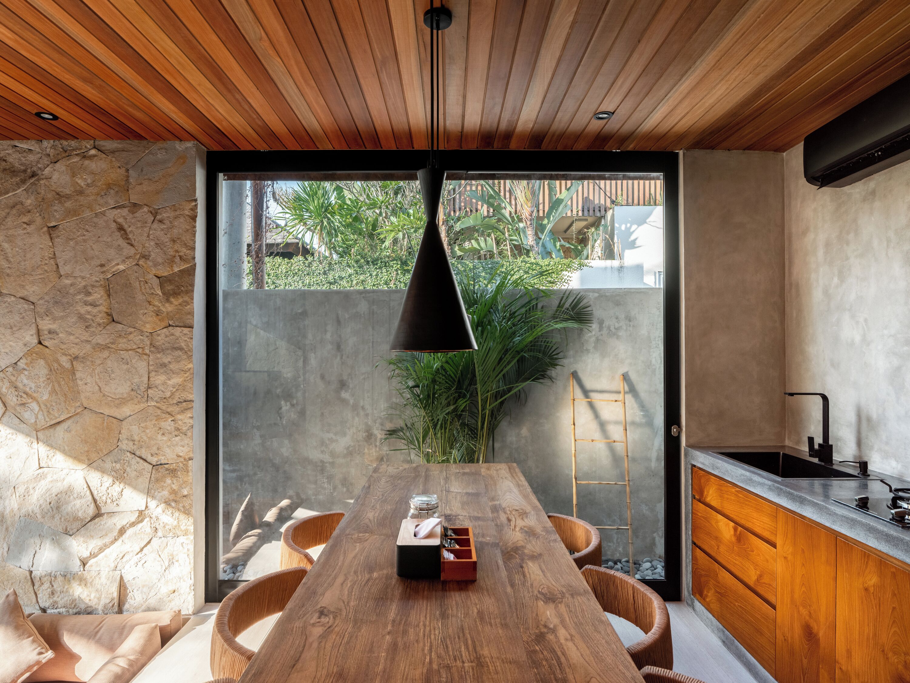 A modern kitchen with a wooden table, stone walls, and a large window overlooking greenery.