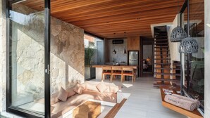 Un interior moderno con pared de piedra, techo de madera y un área de comedor con una cocina al fondo.