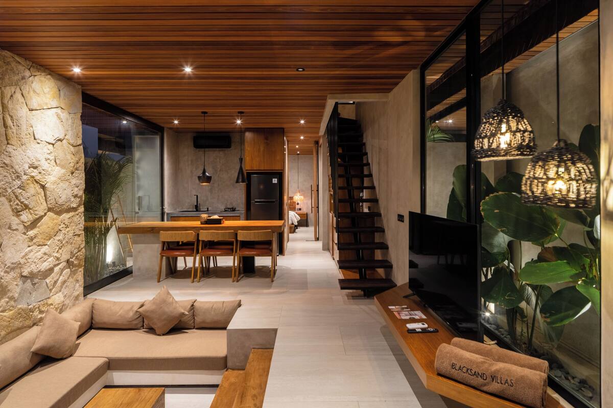 Een modern interieur met een stenen muur, houten plafond, een keuken, een trap en een groot raam met planten.