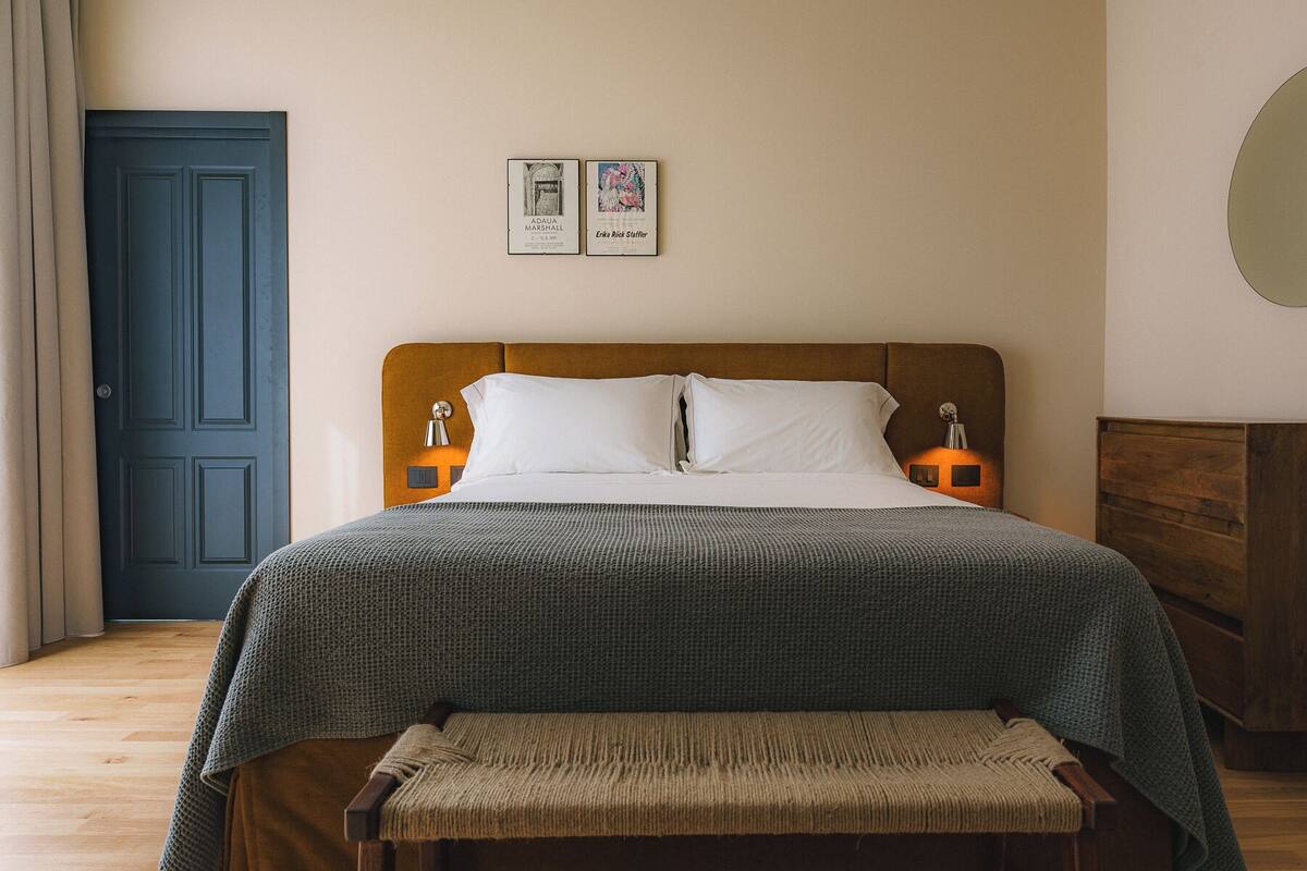 Quarto bem organizado com cama, luminárias de cabeceira, um banco, uma porta e duas fotos emolduradas na parede.