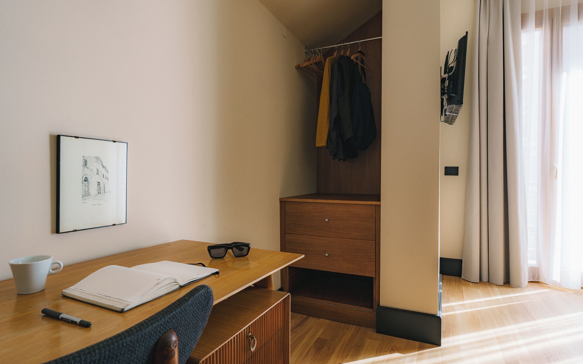 A minimalist room with a wooden desk, a chair, a coat rack with clothes, a small cabinet, and a framed picture on the wall.