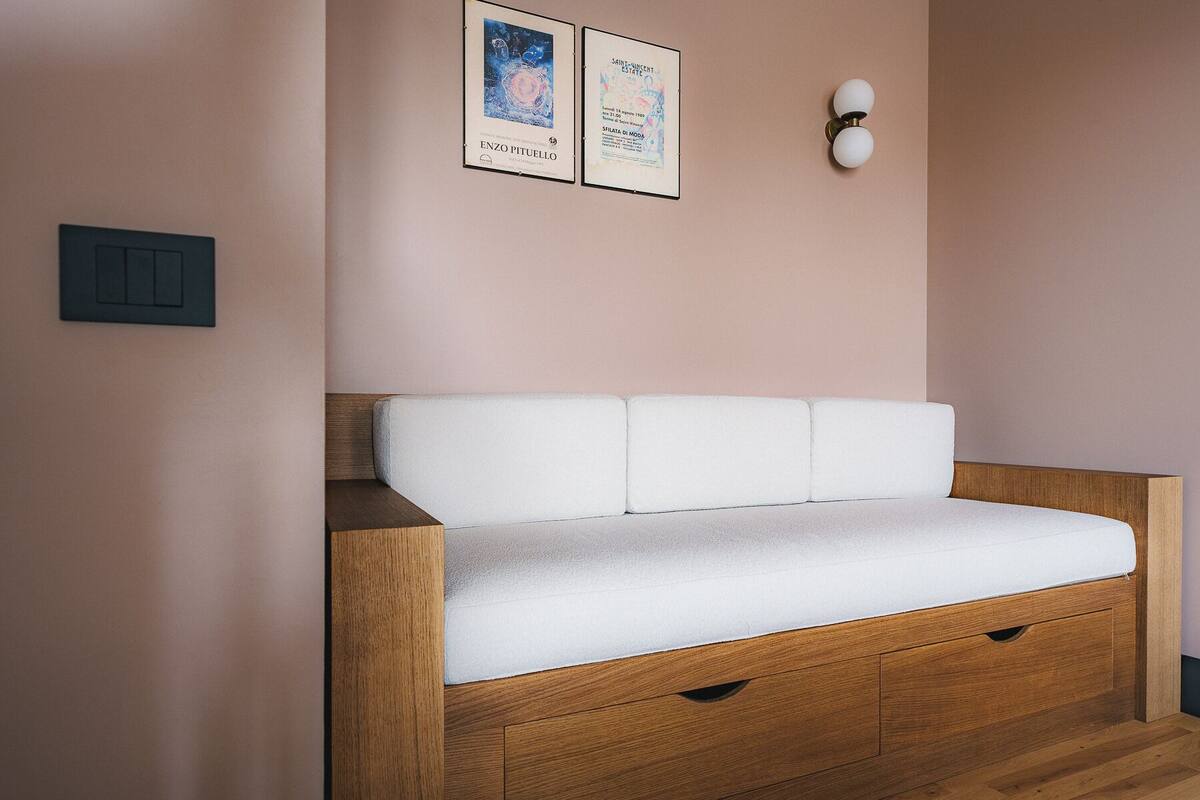 Quarto moderno e minimalista com sofá em estilo banco de madeira, duas obras de arte emolduradas na parede e luminária fixada na parede.