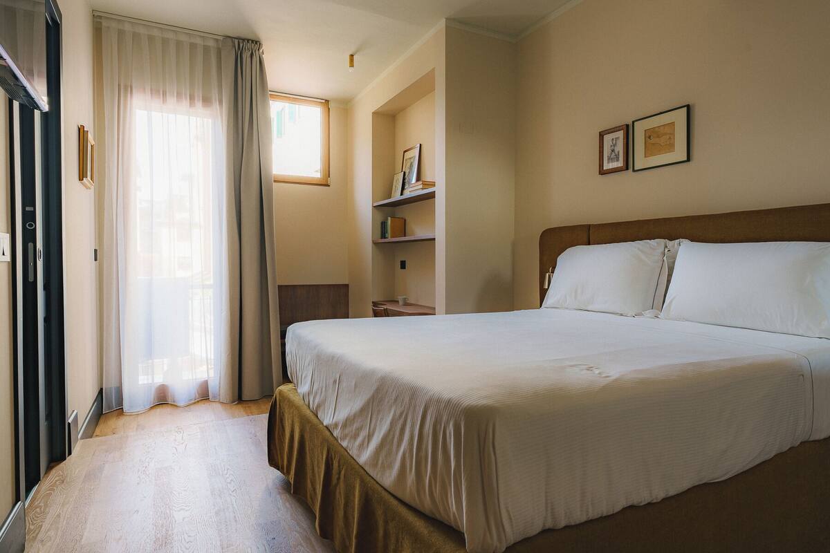 Quarto de hotel com uma cama grande, uma escrivaninha de madeira, uma estante de livros e uma janela com cortinas.