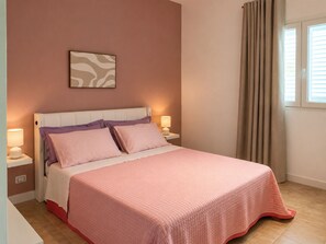 A bedroom with a bed, two bedside lamps, a window with curtains, and a wall-mounted artwork.