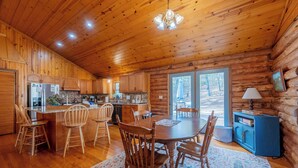 A log cabin interior with a wooden dining table, chairs, a kitchen with wooden cabinets, and a large window.