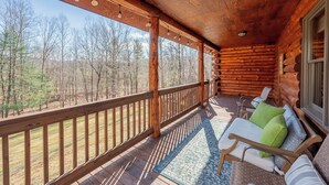 A log cabin with a wooden deck, outdoor seating area with cushions, and a view of the surrounding forest.
