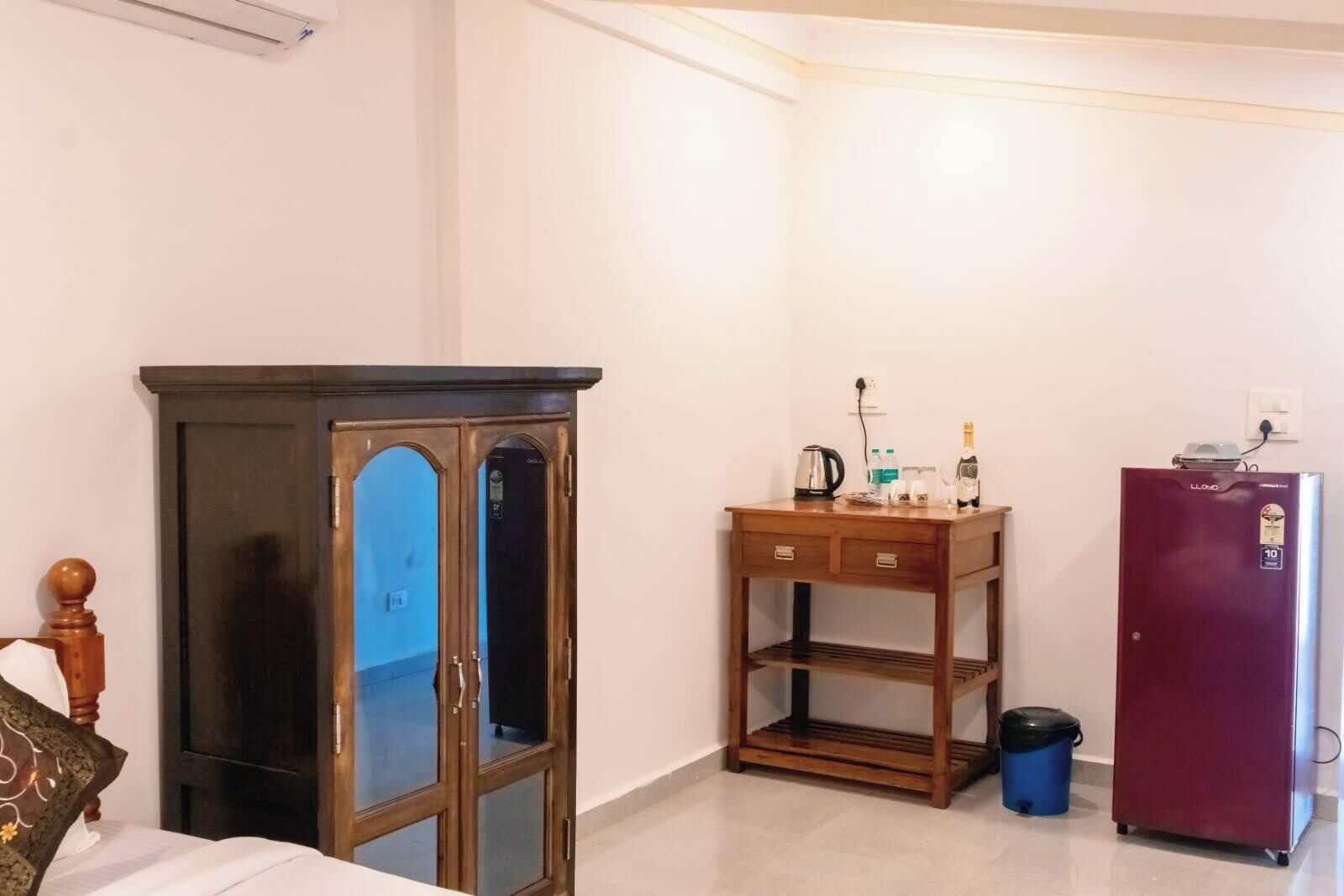 A bedroom with a bed, a wooden cabinet, a small table with a kettle, a refrigerator, and a trash bin.