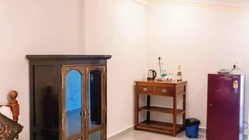 A bedroom with a bed, a wooden cabinet, a small table with a kettle, a refrigerator, and a trash bin.