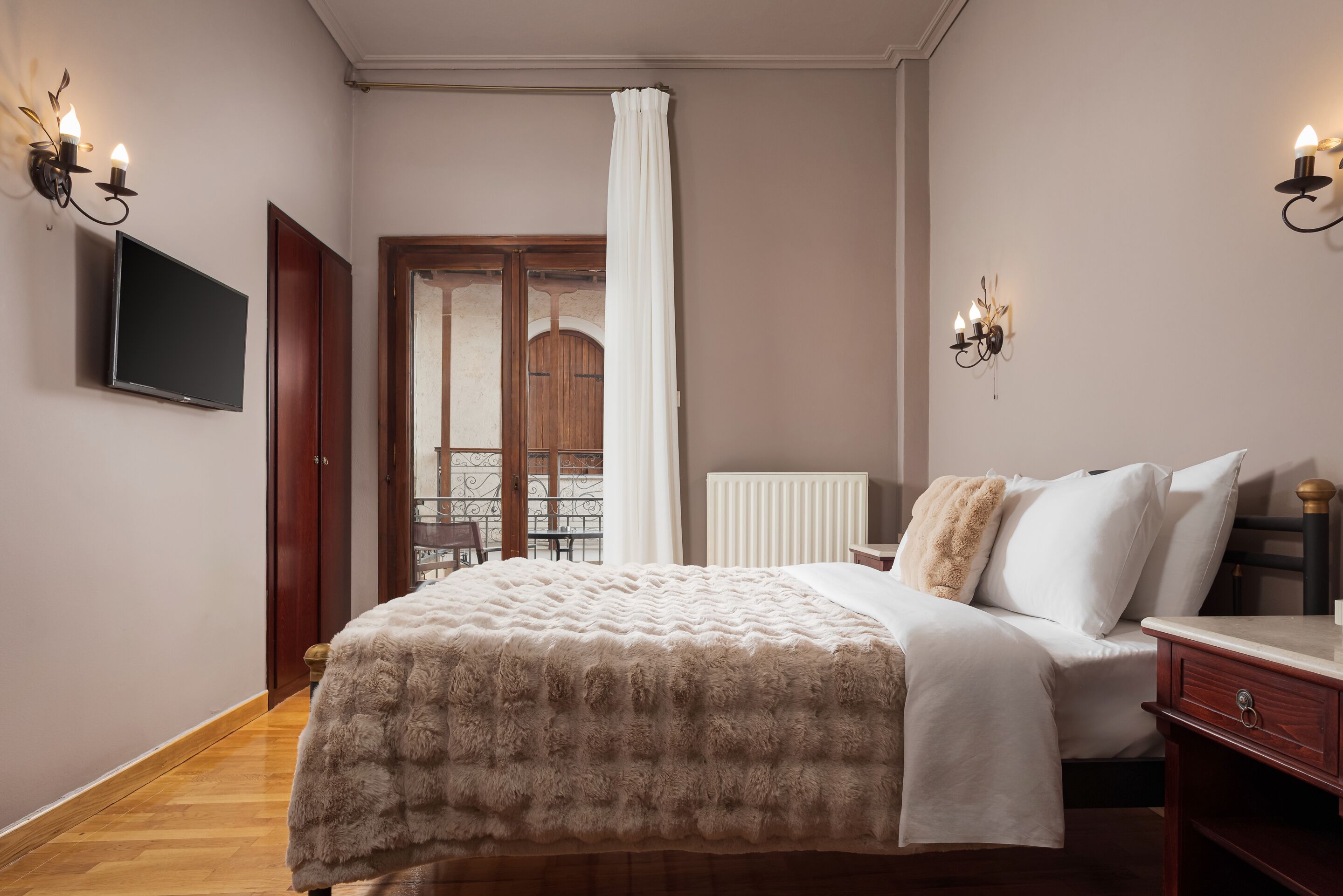 A bedroom with a bed, a TV, a wooden dresser, and a window with a view of a balcony.