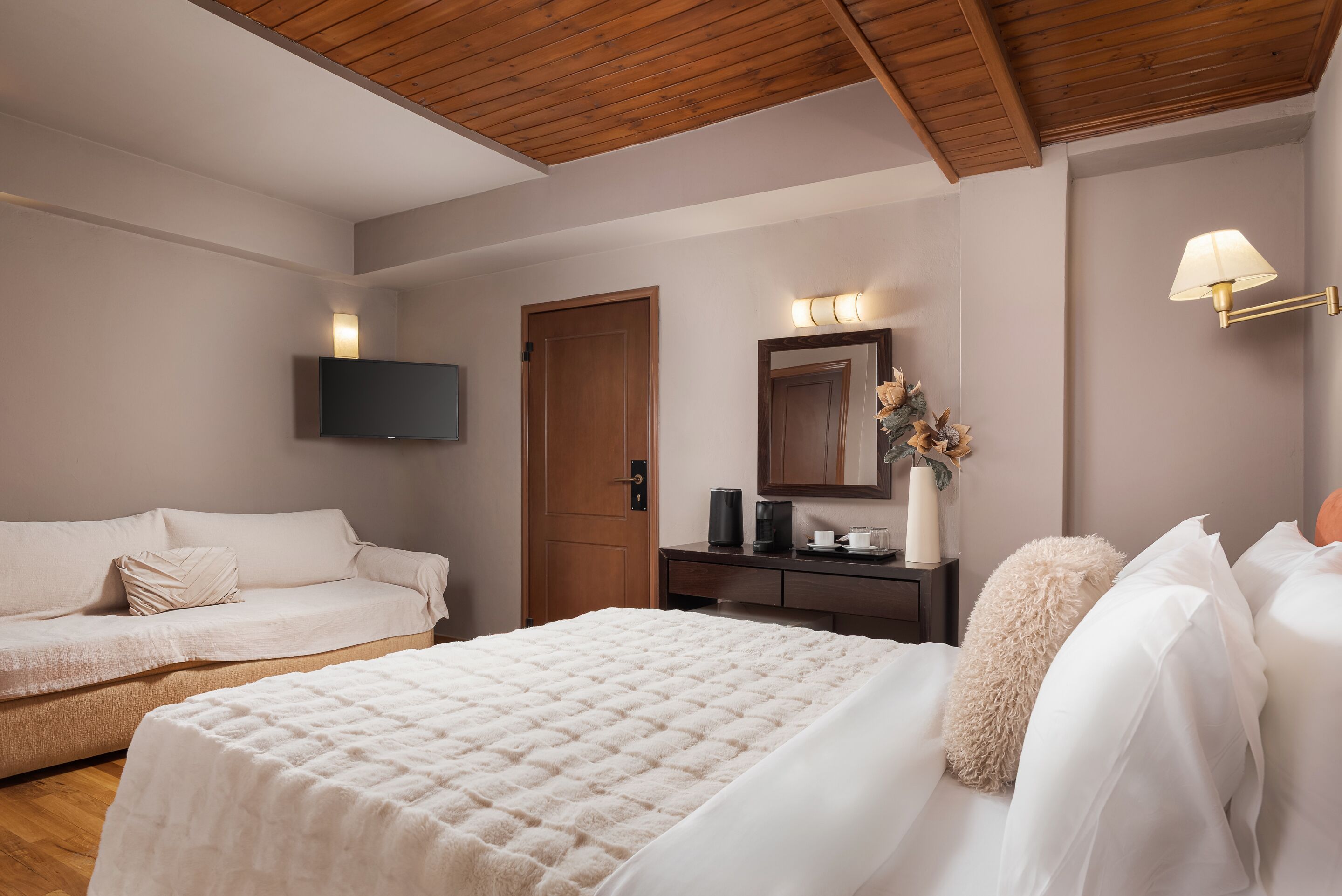 A bedroom with a bed, a sofa, a desk with a lamp, and a TV mounted on the wall.
