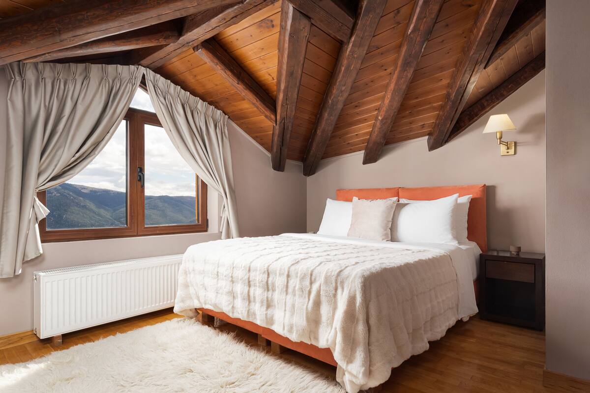 Une chambre avec un plafond en bois, une grande fenêtre avec des rideaux, un lit blanc et une table de chevet avec une lampe.