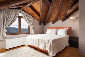 Un dormitorio con techo de madera, un ventanal con cortinas, una cama blanca y una mesita de noche con una lámpara.
